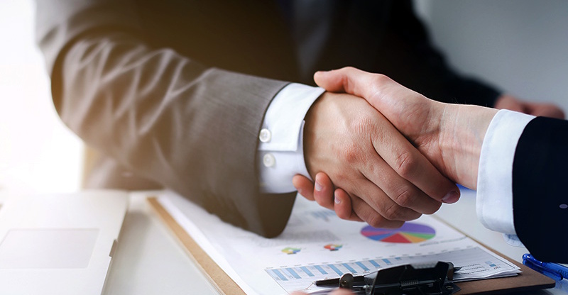 Two people in suits shaking hands over a clipboard with financial charts.