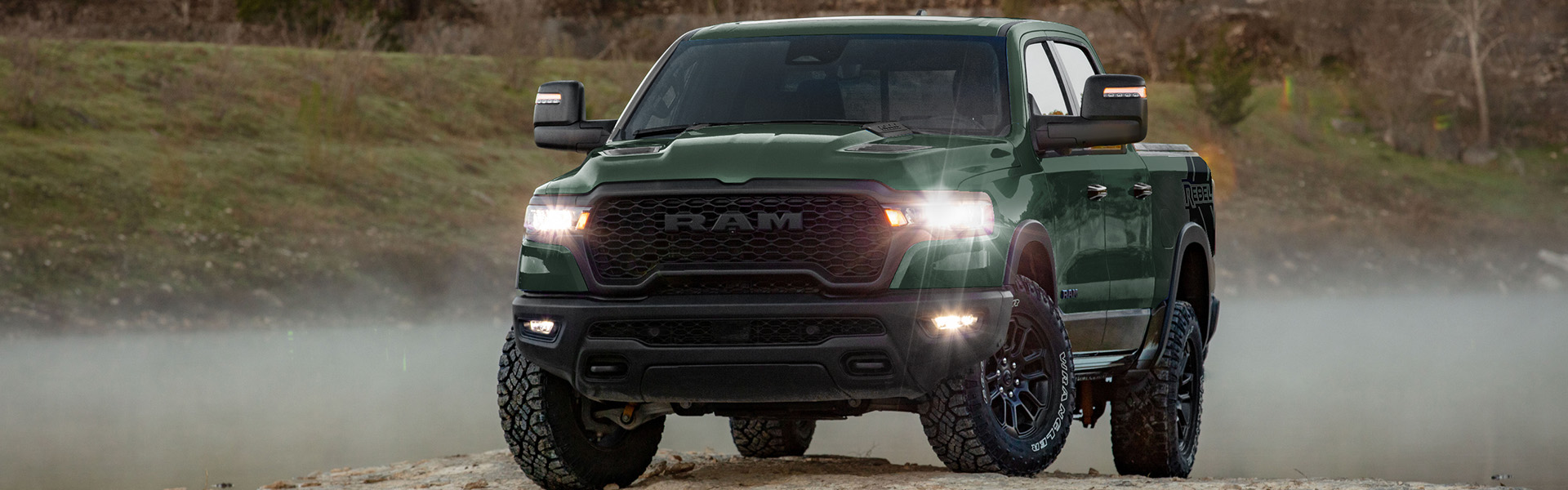 Dark green RAM 1500 Rebel pickup truck parked on a rocky shoreline near a foggy lake.