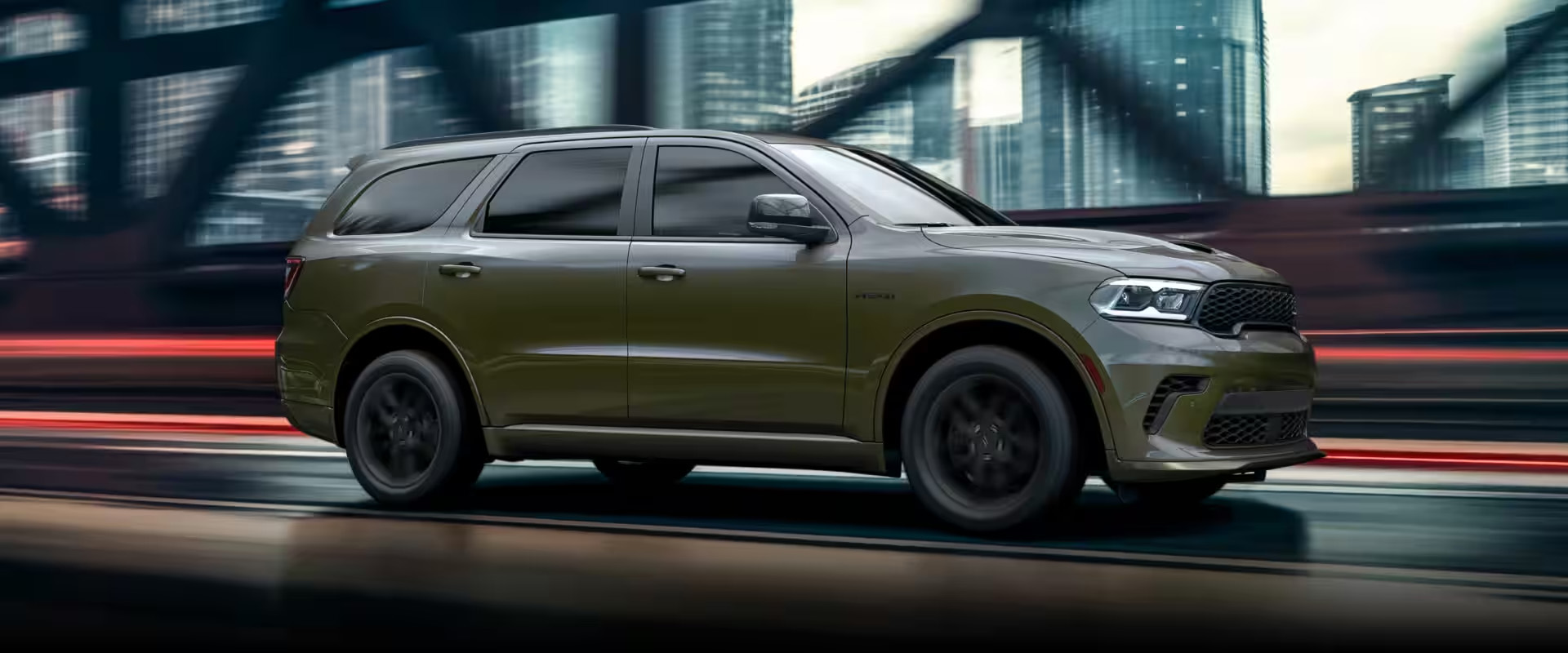 Olive green Dodge Durango SUV driving across a bridge with city skyscrapers in the background.